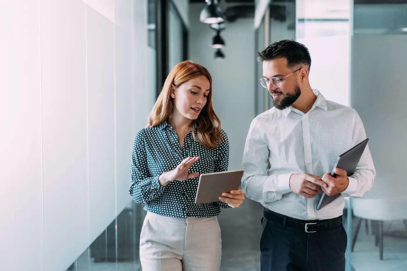 Two smiling colleagues with tablets talking as they walk through a bright office
