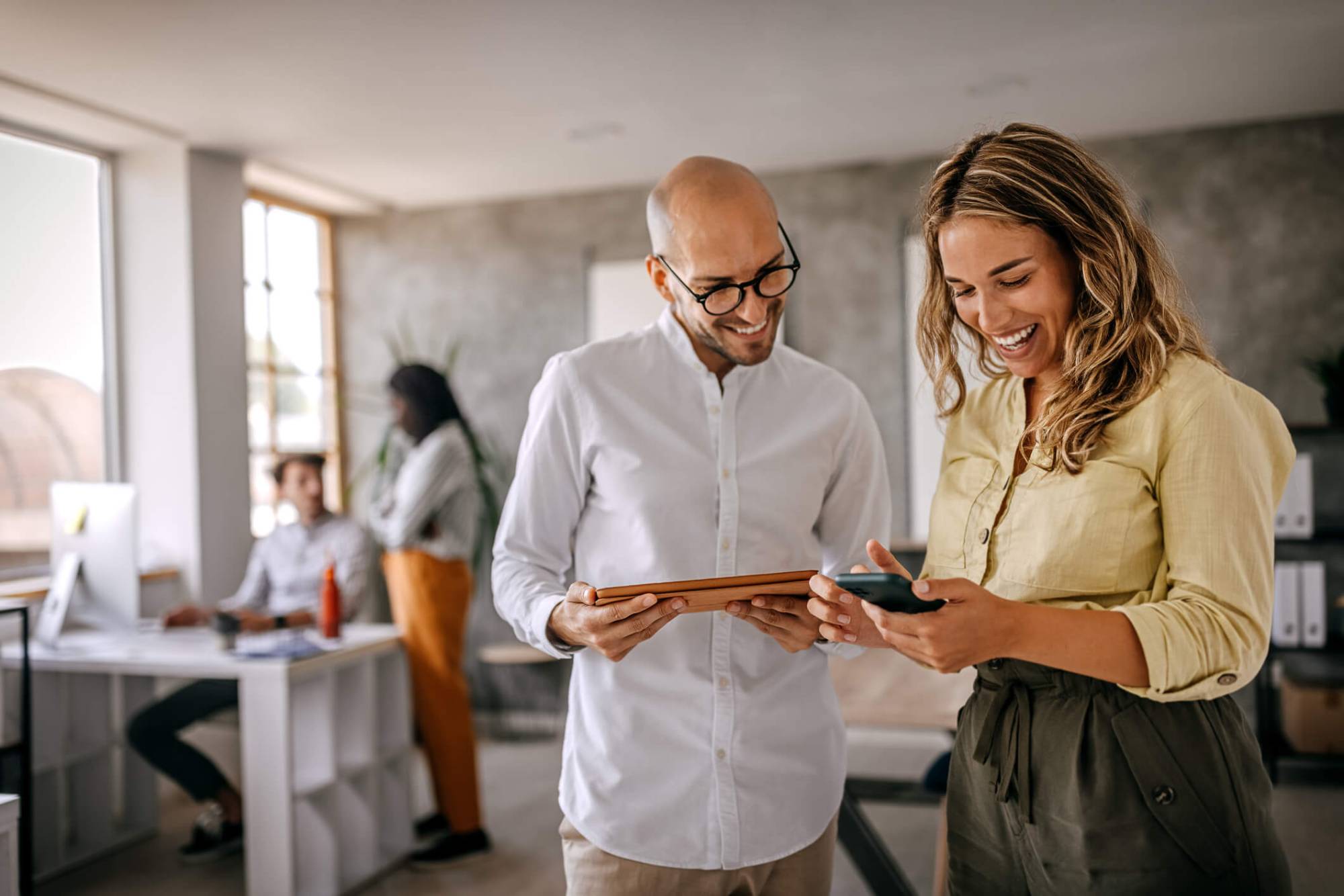 Two colleagues laughing as they look at a phone and a tablet in a bright office