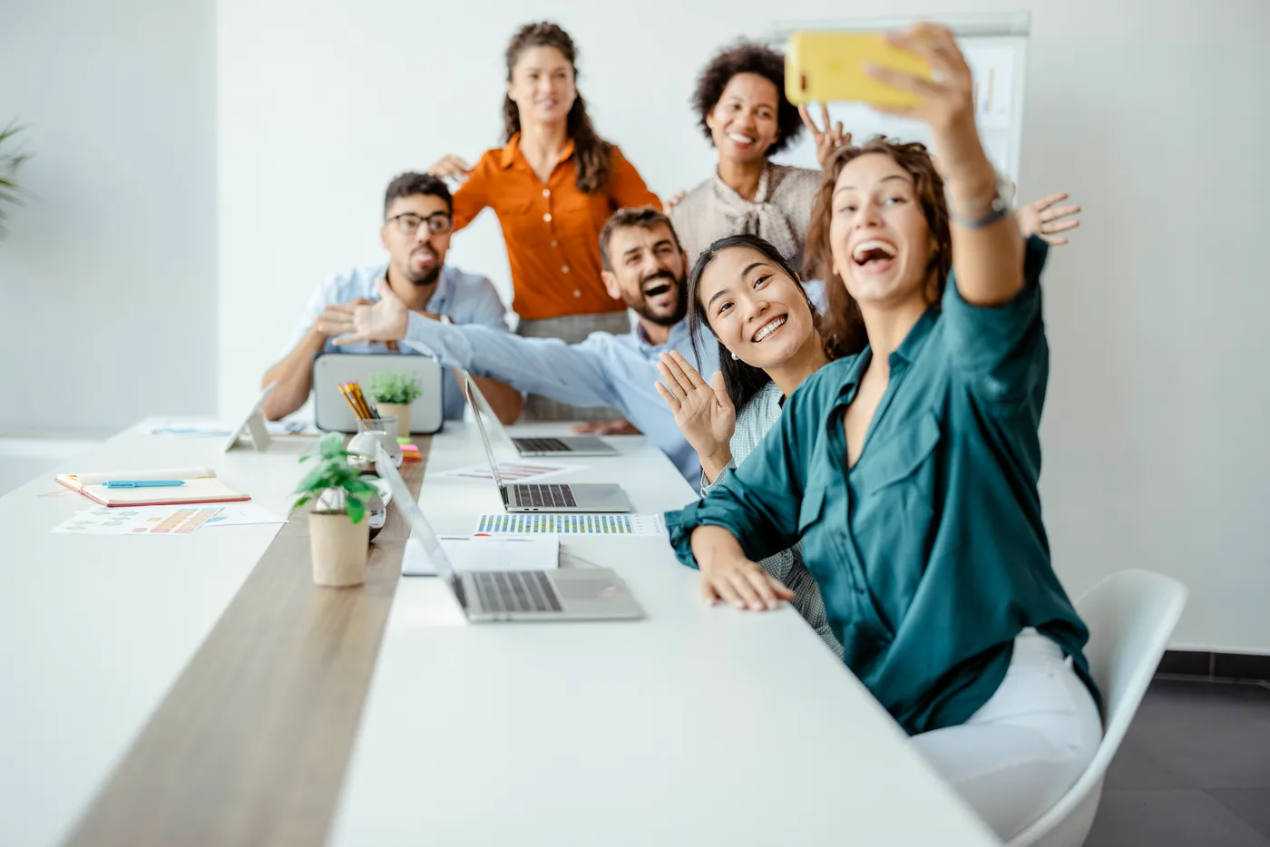 A smiling team taking a group selfie around a meeting table in a bright office