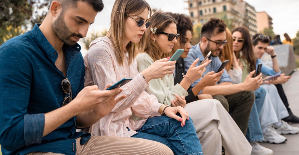 A group of people sitting on a bench looking at their phones